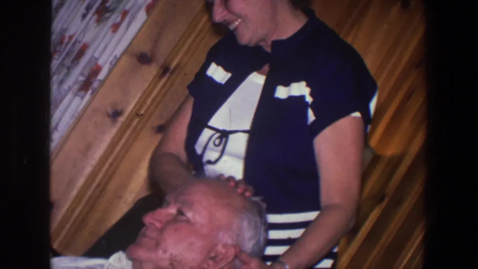 A woman places her hands near the head of a seated elderly man in a warm wood-paneled Saint Paul home interior.