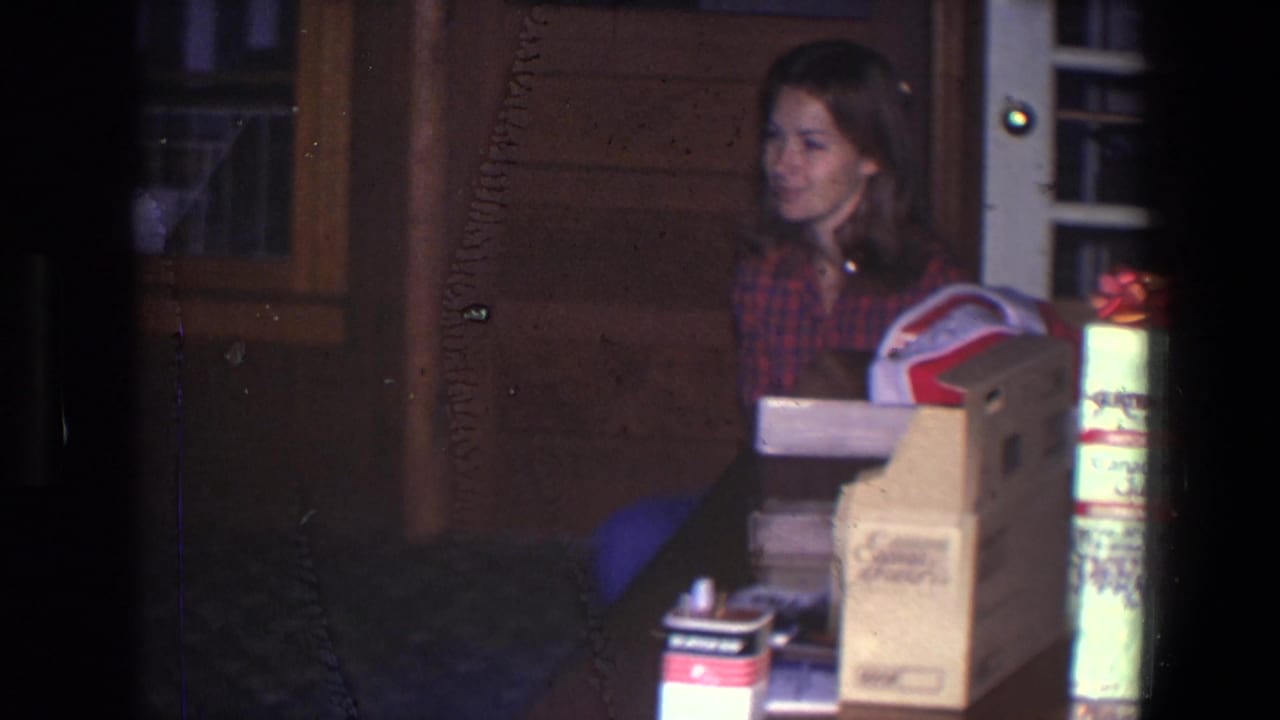 Woman in a red plaid shirt sits indoors beside boxes near a brown door.