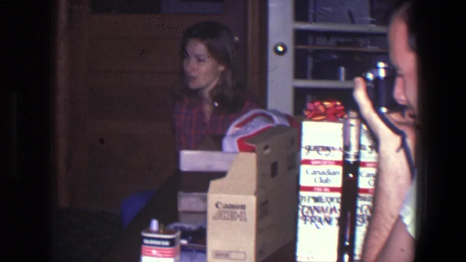 A woman in a plaid shirt sits in a dim Saint Paul interior beside Canon and Canadian Club boxes as another person films her.