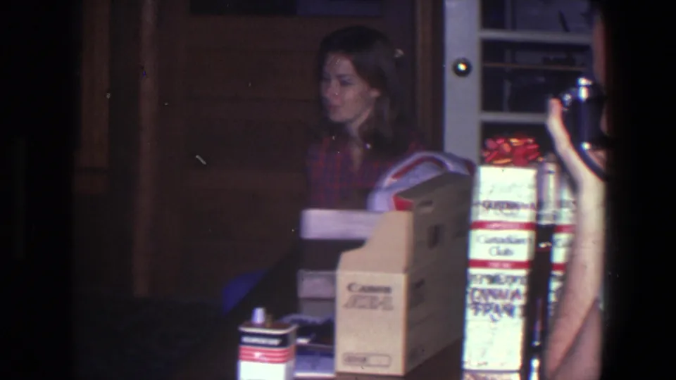 The seated woman looks off-screen in a dim 1971 home interior, with branded boxes and a camera-holding hand in the foreground.