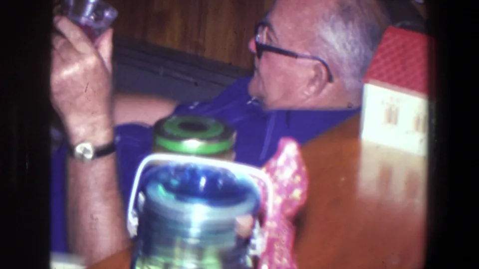 1971 Saint Paul home interior showing a man in a blue shirt and glasses holding a drink near a tabletop with a pitcher and camera packaging.