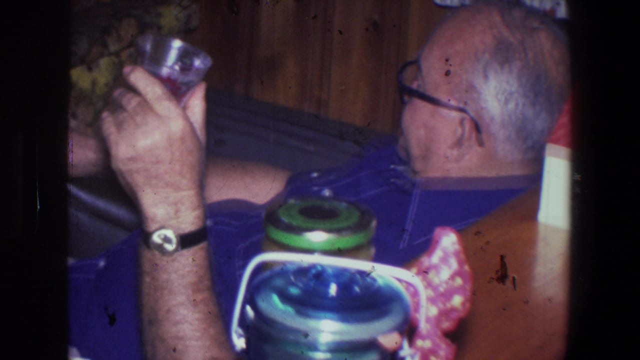 Older man in a blue shirt sits in a Saint Paul home interior in 1971, holding a glass beside a thermos and jars.