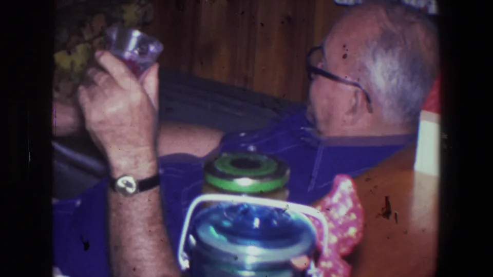 Older man in a blue shirt sits in a Saint Paul home interior in 1971, holding a glass beside a thermos and jars.