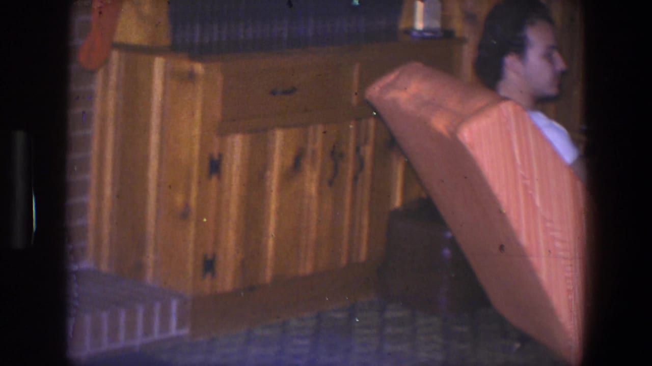 Man in a white shirt seated near wood cabinets and an upholstered chair.