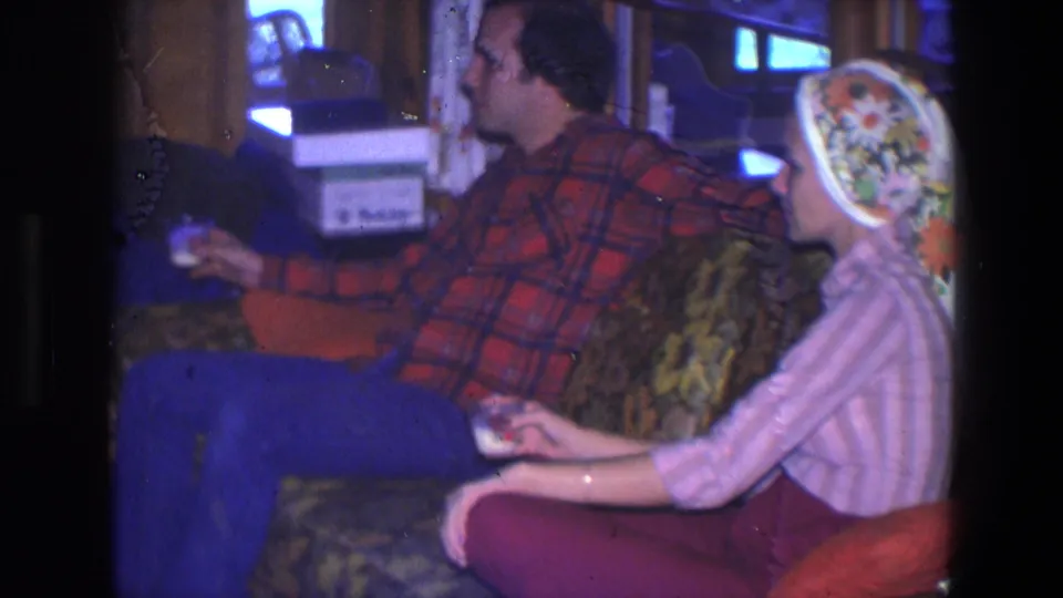 1971 Saint Paul home interior frame with a man in a red plaid shirt and a woman in a pink striped top seated on a patterned sofa.