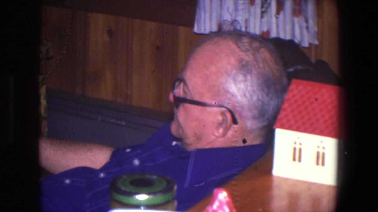 Older man in glasses and a blue shirt sits in a Saint Paul paneled den, with curtains, a tabletop, and a small model house nearby.