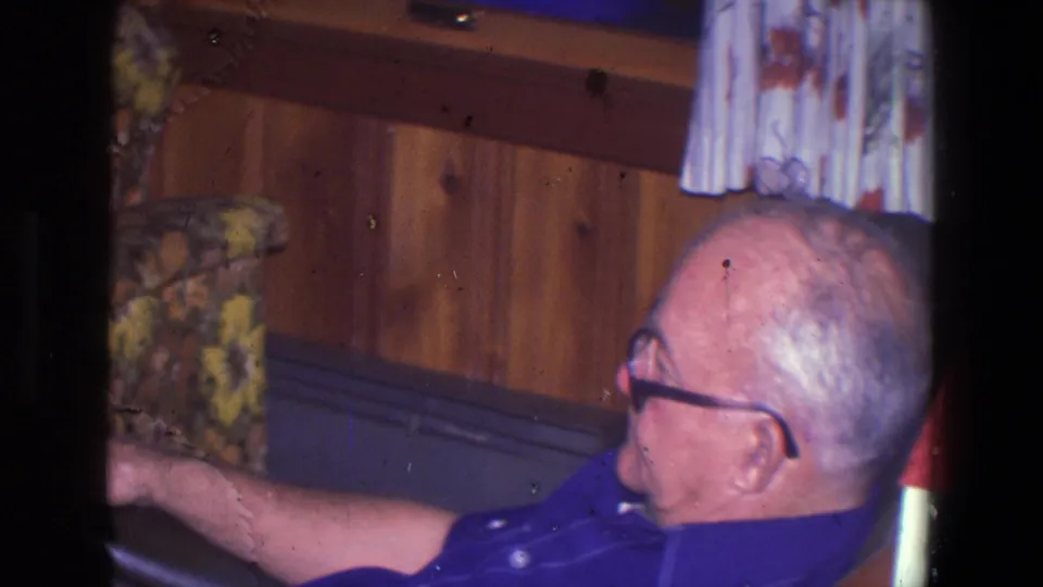 Older man in glasses appears in profile in the 1971 Saint Paul interior, seated near wood paneling and patterned furnishings.