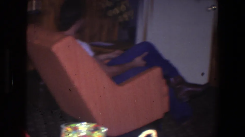 Younger man sits in an orange recliner inside the Saint Paul den, with wood paneling, a curtain, and tabletop objects partly visible.