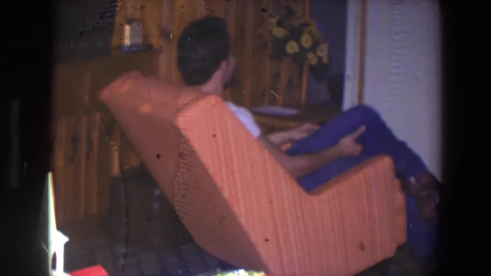 Younger man is seen from behind in an orange recliner in the 1971 Saint Paul den, with wood paneling, floral curtains, and a small model house visible.