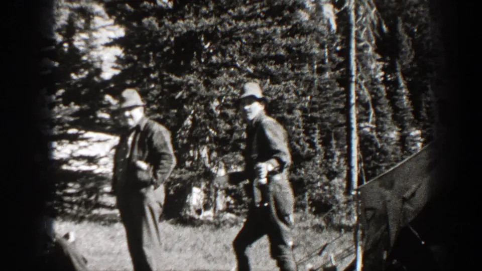 Two men in hats and outdoor clothing walk through a forested California mountain area, with part of a horse visible nearby.
