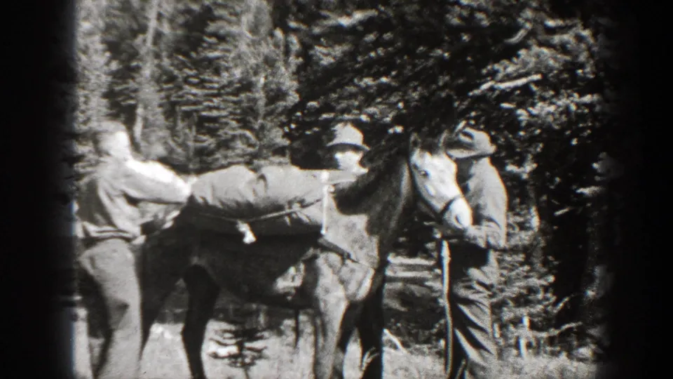 Two men in period outdoor clothing stand beside a horse in the woods, appearing to secure or inspect its tack.