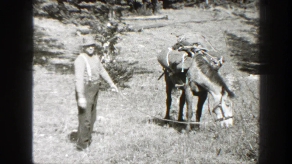 1938 California 8mm frame showing a man in hat and suspenders near a loaded pack mule in a mountain clearing.