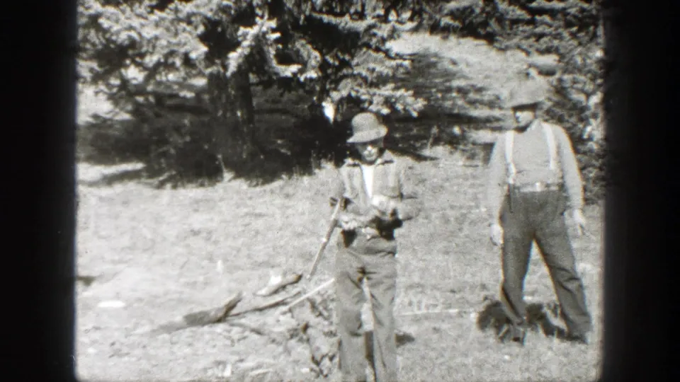 Two men in period outdoor clothing stand near branches and gear at a California mountain campsite.