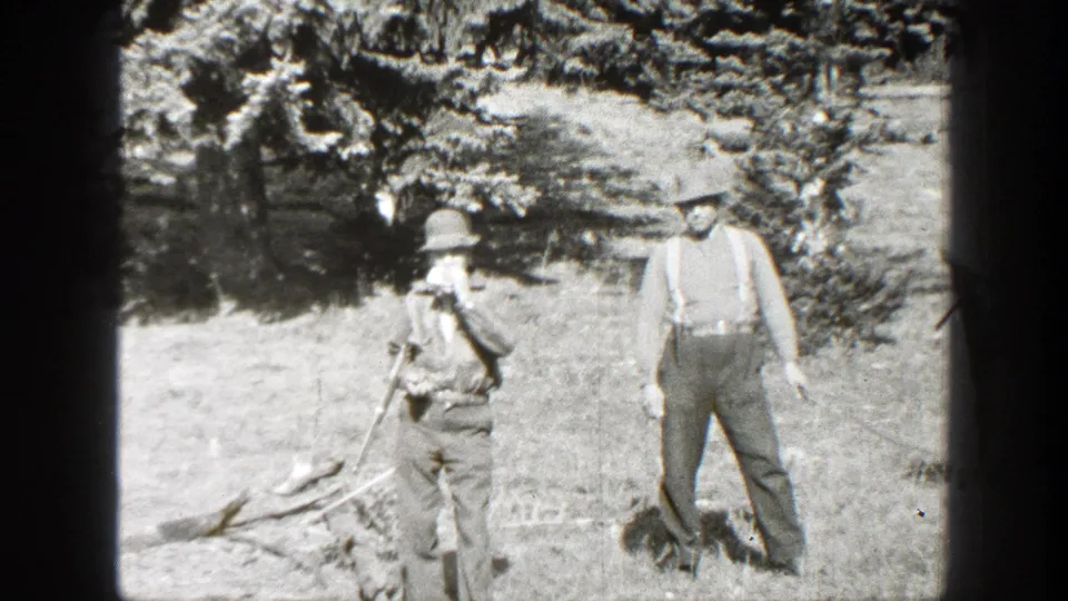 1930s home-movie frame of two men in hats and work clothes standing in a California campsite clearing.