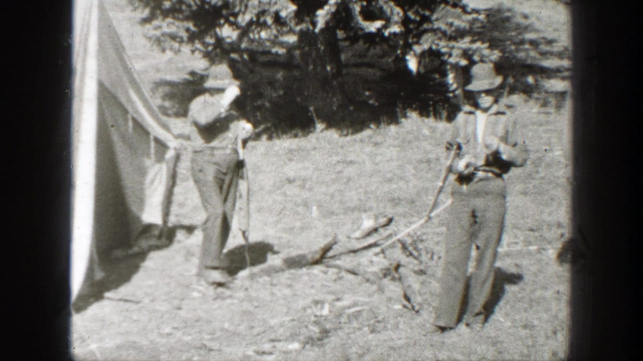 Two adult men in period outdoor clothing stand by a canvas tent in a California wooded campsite, 1938 home-movie footage.