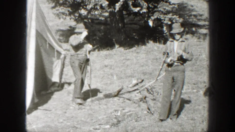 Two adult men in period outdoor clothing stand by a canvas tent in a California wooded campsite, 1938 home-movie footage.