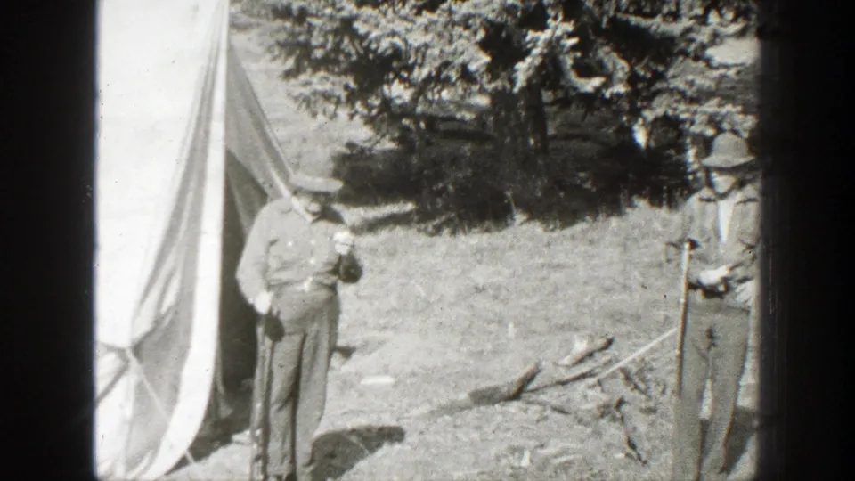 Two adult men in hats stand beside a canvas tent and fallen branches in the 1938 California campsite footage.