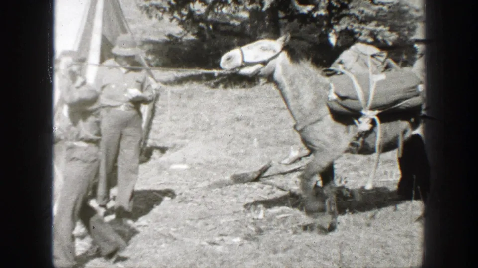 1938 California home-movie frame showing a man with a rifle near a loaded pack mule at a rustic campsite.
