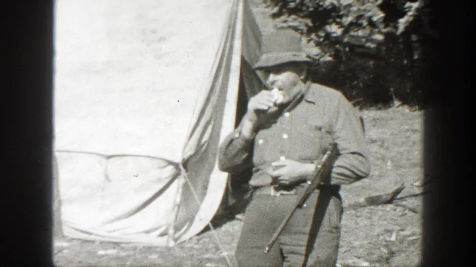 1938 California frame showing a man in period outdoor clothing holding a rifle near a canvas tent.