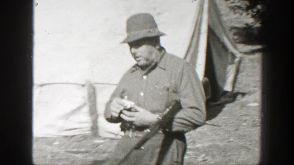 1938 California 8mm frame of a man in military-style clothing smoking a cigar beside a tent with a rifle held at his side.