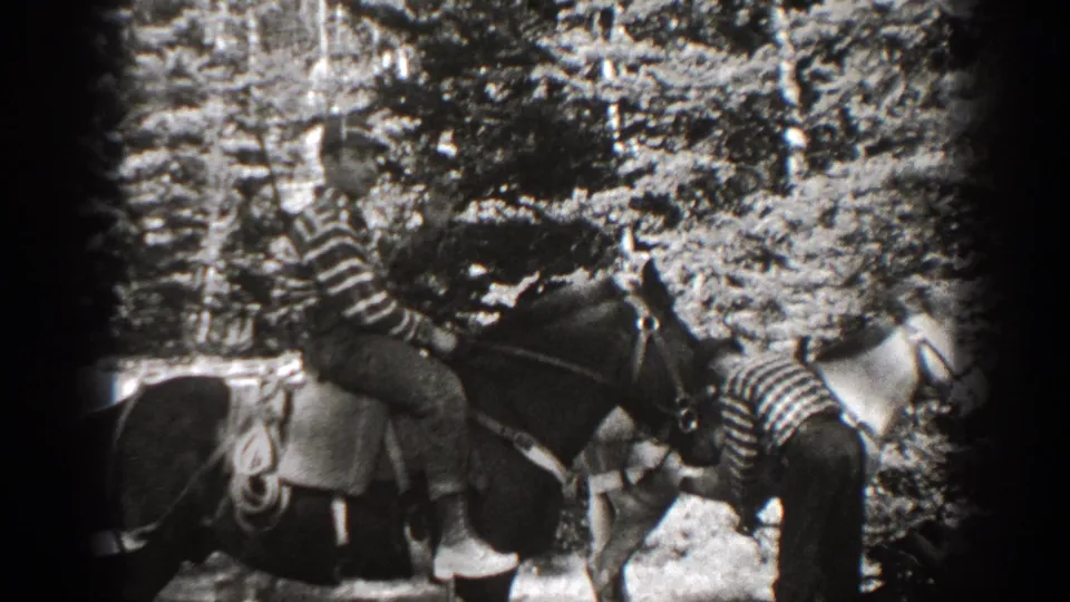 1938 California 8mm frame of a striped-shirt rider on a saddled horse with another person bending near a second horse in a wooded area.