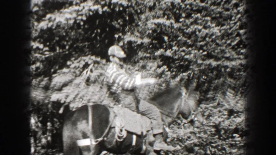 1938 California home-movie frame of an adult man in a striped shirt and cap riding a horse through forest underbrush.