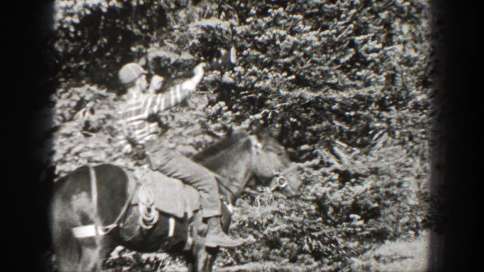 Black-and-white 8mm frame of a man on horseback lifting a rifle in a wooded California setting with dense shrubs behind him.
