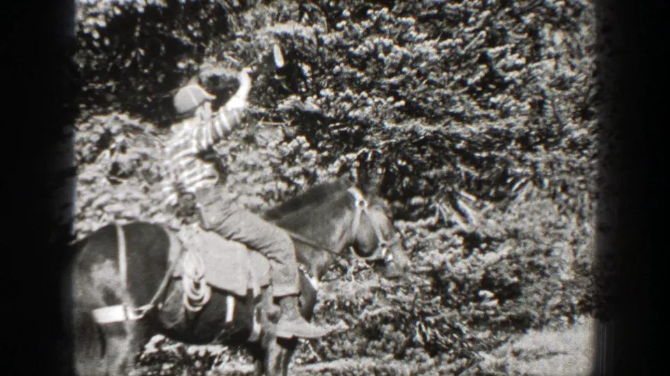 1938 California home-movie frame showing a rider holding a rifle high from the saddle against dense forest vegetation.