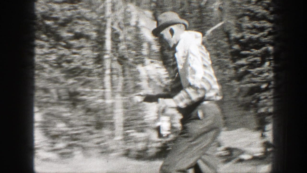 Man in a hat and striped shirt walks through trees carrying a bottle or container.