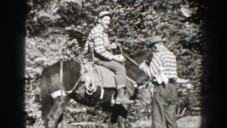A mounted man sits on a saddled horse as another man in a striped shirt stands beside him in a wooded California clearing.