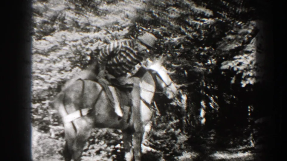 In 1938 California home-movie footage, a man in a striped shirt and hat mounts a saddled horse in a wooded area.