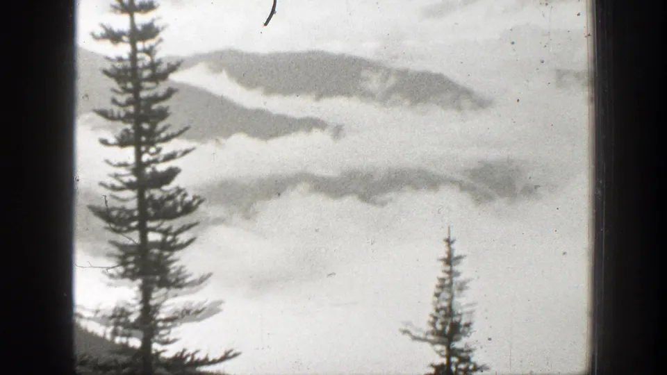 A black-and-white 1938 California mountain view with evergreen silhouettes, low clouds, and distant ridgelines behind a window frame.