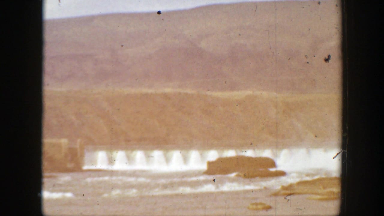 Faded 8mm view of water spilling over a dam with hills behind it.