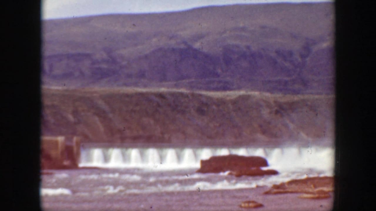 Dam spillway with white water and a dark object in the river below hills.