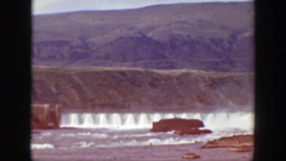 1938 California home-movie frame of water pouring over a dam, with a dark rock or structure in the river and rugged hills beyond.