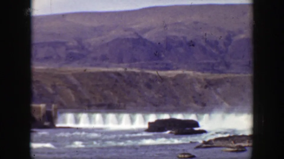 1938 California 8mm frame showing the spillway from a slightly different angle, with foreground water and hazy mountain slopes in back.