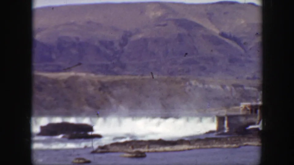 1938 California 8mm frame showing the dam and river from another angle, with rocks in the foreground and a small structure near the right edge.
