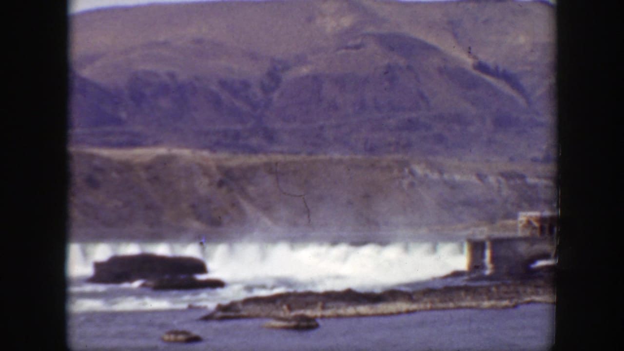 1938 California 8mm frame showing a dam releasing water into a river, with rocky banks and mountains in the background.
