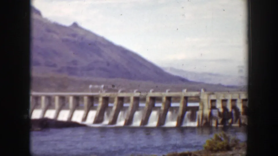 1938 California 8mm frame showing the dam from farther right, with repeated spillway bays and mountains beyond.