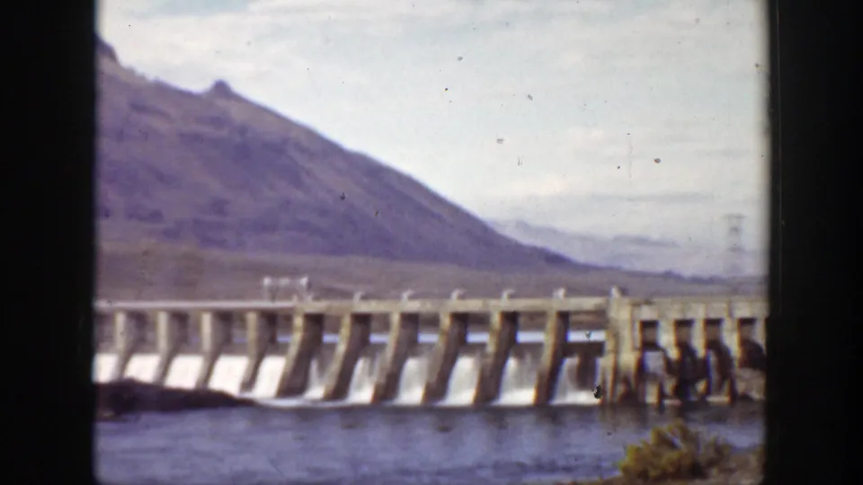 1938 California 8mm frame with the dam, water below, and a broad mountain slope under a pale sky.