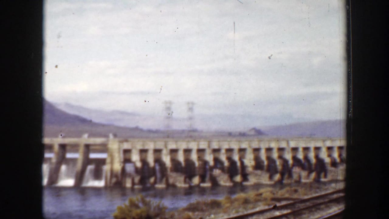 Concrete dam with water gates, mountains, rail tracks, and small distant figures.