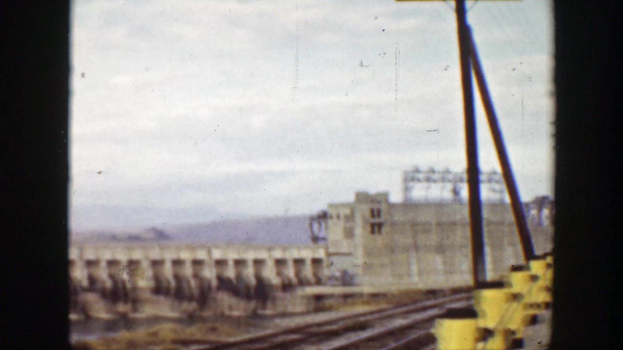 Dam section with industrial building, rail tracks, mountains, and yellow roadside bollard.