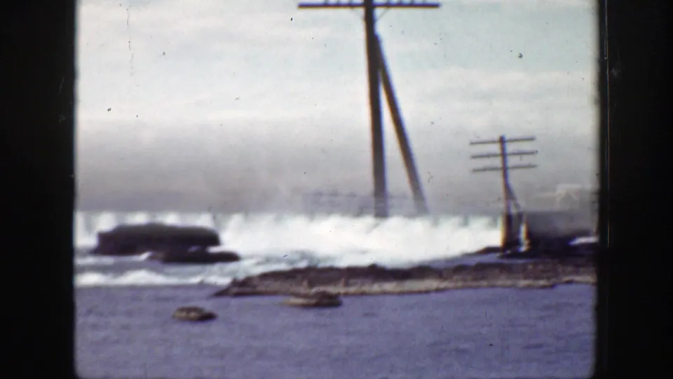 1938 California 8mm frame showing turbulent water by rocks, leaning telephone poles, a utility pole, and hazy mountains in the background.