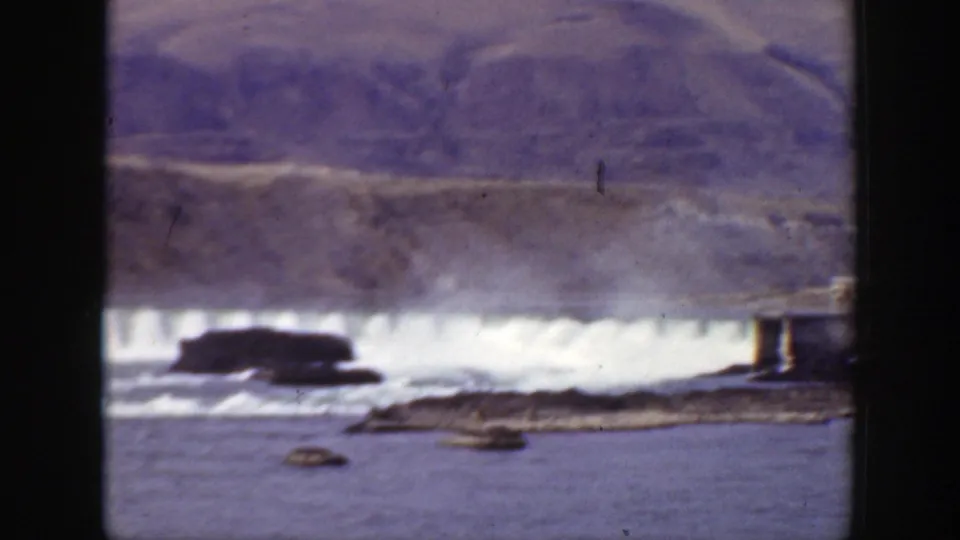 1938 California 8mm frame of foaming waterside flow below a mountain slope, with rocks and a telephone pole near the edge of the water.