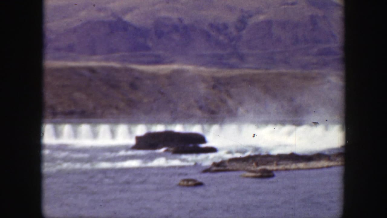 1938 California home-movie frame showing a dam spillway, white water, rocky foreground, and barren hills in soft 8mm color.