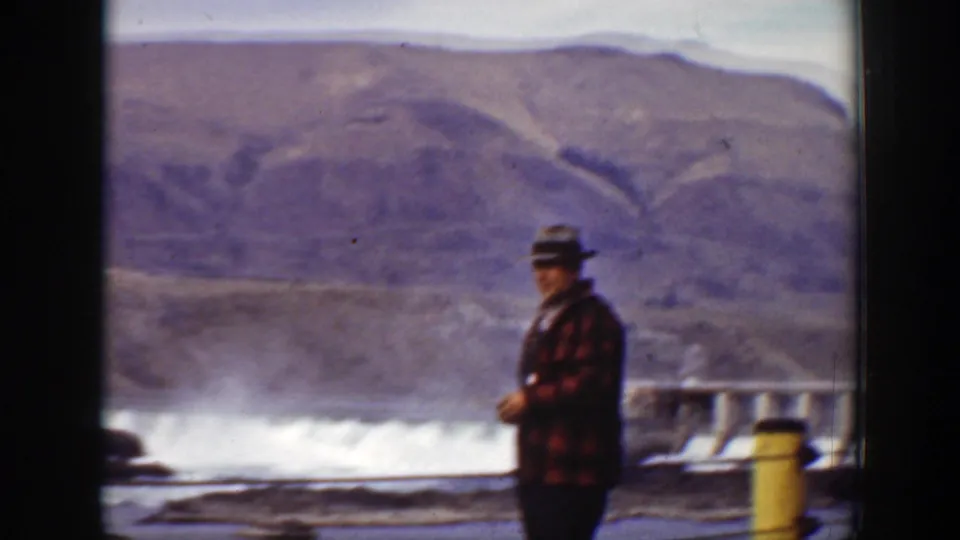 An adult man in a hat, checkered jacket, and overalls stands near the dam spillway in the supplied 1938 California home-movie footage.