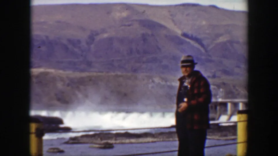 A hat-wearing adult man stands beside yellow posts or railings, with a California dam spillway and dry hills behind him.