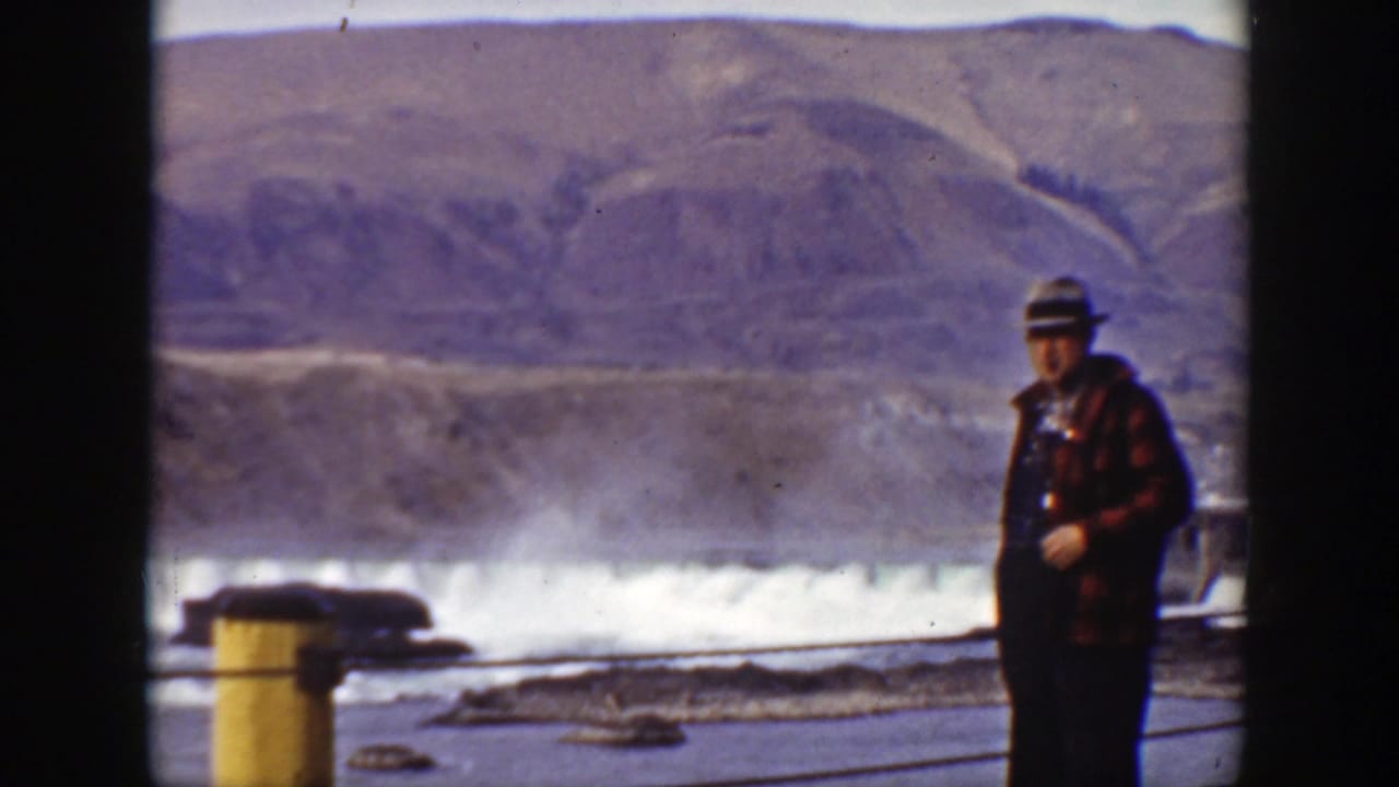 Man stands farther right beside the dam, with rushing water and mountains behind him.