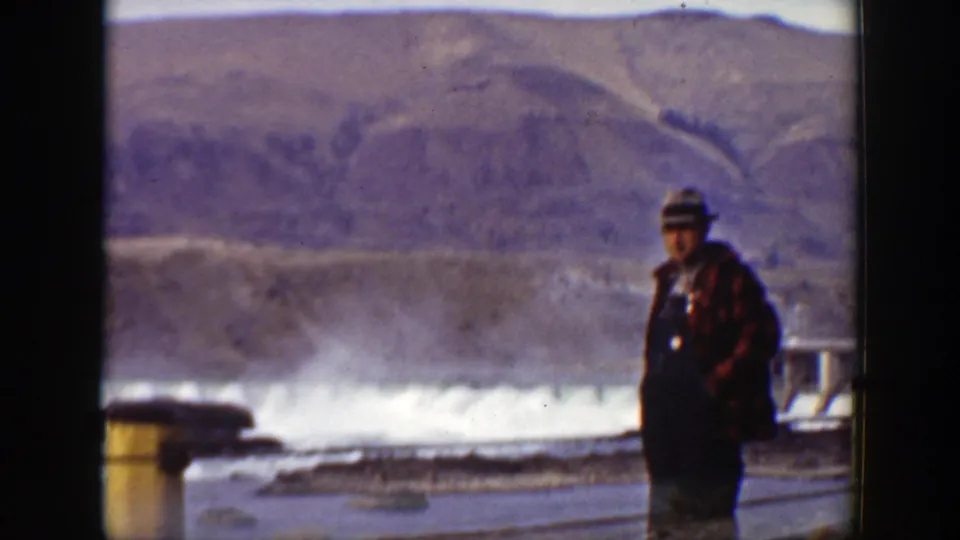A visitor in a gray hat and plaid jacket stands in front of the dam release, with white spray and hills behind him.