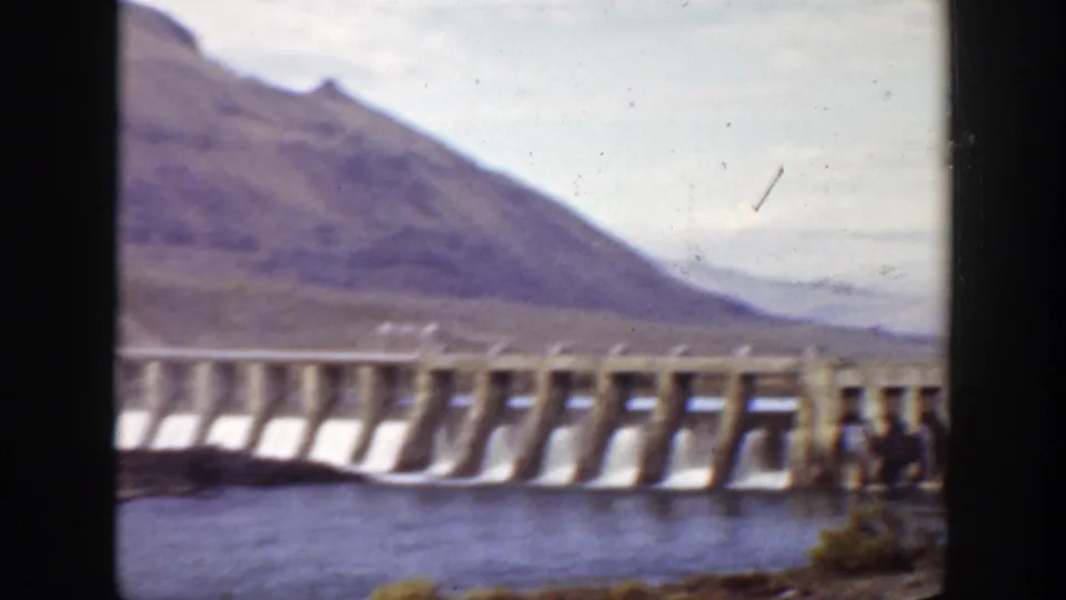 The final frame shifts toward the concrete dam structure, showing water below, mountains beyond, and part of a person at the right edge.